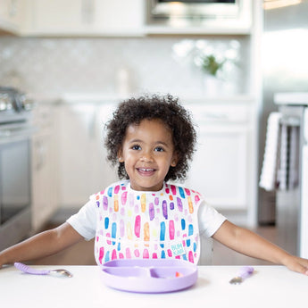 A toddler smiles in a bright kitchen, wearing a colorful bib at a white table with a Bumkins Silicone Grip Dish: Lavender and utensils.