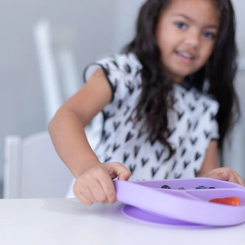 A child in a black & white bib lifts a Bumkins Silicone Grip Dish: Lavender filled with berries on a white table.