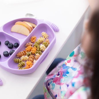 A child in a colorful bib enjoys pasta, apple slices, and blueberries from a Bumkins Silicone Grip Dish: Lavender at a white table.