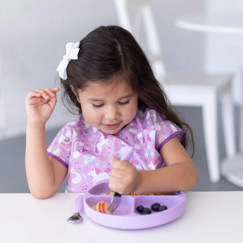 A young girl enjoys a meal from a Bumkins Silicone Grip Dish: Lavender at a white table, wearing a purple unicorn bib and a white bow.