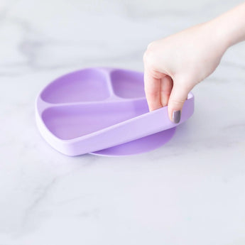 A hand holds a Bumkins Silicone Grip Dish: Lavender, demonstrating the suction cup bottom on a marble surface.