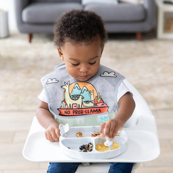 A toddler wearing a llama bib enjoys fruit and snacks from a Bumkins Silicone Grip Dish: Marble with utensils while seated in a high chair.
