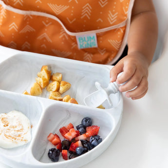 Close-up of a child in an orange bib eating pancakes, fruit, and yogurt from a Bumkins Silicone Grip Dish: Marble with a matching spoon.