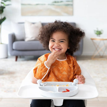 A curly-haired toddler in an orange bib sits happily in a highchair, eating from a Bumkins Silicone Grip Dish: Marble in a cozy living room.
