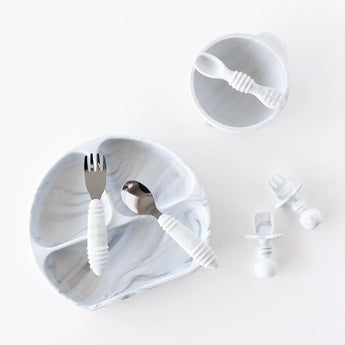 Utensils sit on top of a Bumkins Silicone Grip Dish: Marble on a white background, with toddler utensils and a bowl & spoon set nearby.
