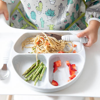 Close-up of a child in a cactus bib enjoying spaghetti, asparagus, and strawberries from a Bumkins Silicone Grip Dish: Marble with a fork.