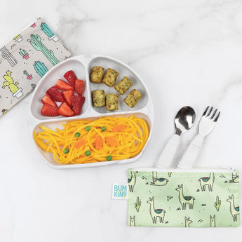 Bumkins Silicone Grip Dish: Marble holds pasta, fruit, and veggies on a marble surface, with green and gray pouches and utensils nearby.