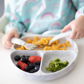 Close-up of a child in rainbow bib eating pasta, berries, and broccoli from a Bumkins Silicone Grip Dish: Marble using a fork and spoon.