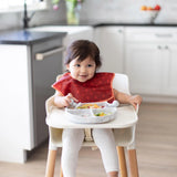 A toddler enjoys food from a Bumkins Silicone Grip Dish: Marble with toddler utensils, seated in a highchair wearing a red bib.