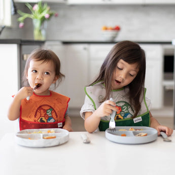 Two kids in bibs dine happily at a white table in a kitchen; one eats from a Bumkins Silicone Grip Dish: Marble.