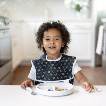A child with curly hair in a black bib grins in a bright kitchen, sitting with food in a Bumkins Silicone Grip Dish: Marble on a white table.