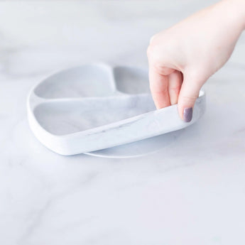 A hand holds a Bumkins Silicone Grip Dish: Marble, demonstrating the suction cup bottom on a marble surface.