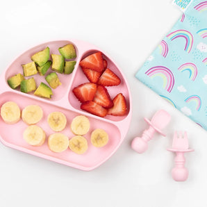 Bumkins Silicone Grip Dish: Pink with avocado and fruit on a white background, with pink toddler utensils and a rainbow-print pouch nearby.