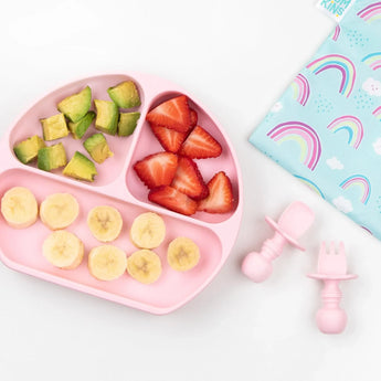 Bumkins Silicone Grip Dish: Pink with avocado and fruit on a white background, with pink toddler utensils and a rainbow-print pouch nearby.