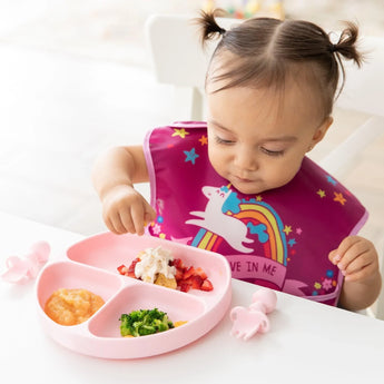 A toddler wearing a unicorn bib eats broccoli, fruit, & applesauce from a Bumkins Silicone Grip Dish: Pink at a white table.