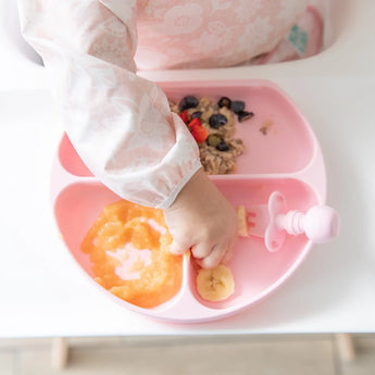 Close-up of a toddler reaching for food from a Bumkins Silicone Grip Dish: Pink on a high chair tray.