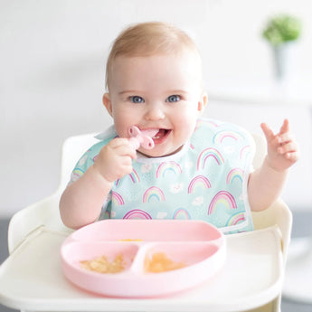 A smiling baby in a high chair wears a rainbow-print bib and holds a pink toddler spoon with a Bumkins Silicone Grip Dish: Pink on the tray.