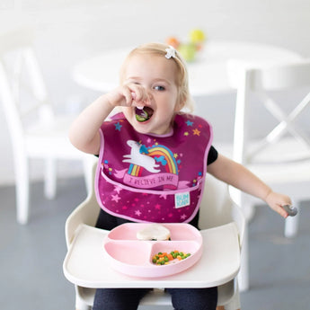 A toddler in a high chair, wearing a unicorn bib, enjoys self-feeding from a Bumkins Silicone Grip Dish: Pink with pink utensils.