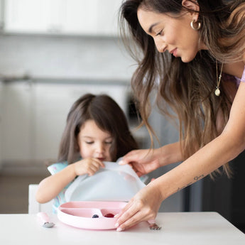 In a bright kitchen, a woman aids a child by removing a lid from a Bumkins Silicone Grip Dish: Pink on a white table.