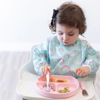 A toddler in a high chair, wearing a rainbow bib, uses pink utensils to eat fruit and pasta from a Bumkins Silicone Grip Dish: Pink.