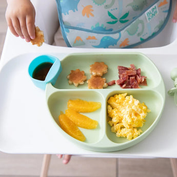 Close-up of a child in a high chair with a Bumkins Silicone Grip Dish: Sage filled with breakfast foods and a blue cup of syrup on the tray.