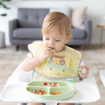 A toddler wearing a camping-themed bib enjoys a meal in a high chair from Bumkins Silicone Grip Dish: Sage with matching utensils.
