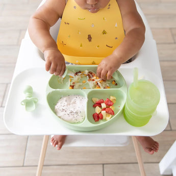 A baby in a high chair enjoys yogurt, chopped fruit, and hash browns from a Bumkins Silicone Grip Dish: Sage with utensils and a green cup.