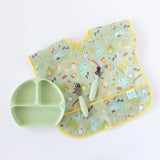 Bumkins Silicone Grip Dish: Sage sits near a camping-themed bib and green utensils on a white background.