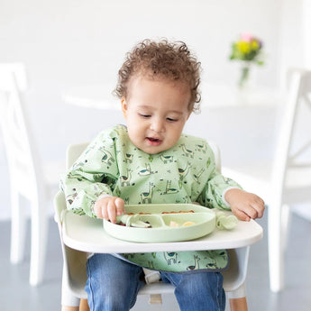 A curly-haired toddler in a high chair wearing a green bib eats from a Bumkins Silicone Grip Dish: Sage with matching utensils.
