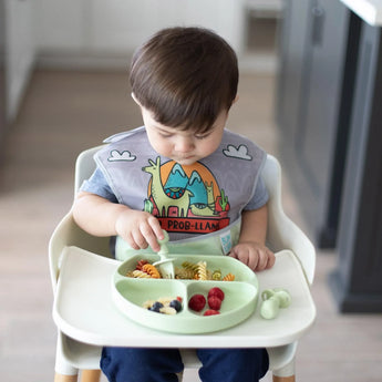 A toddler in a llama bib sits in a high chair, eating pasta and berries from a Bumkins Silicone Grip Dish: Sage with matching utensils.