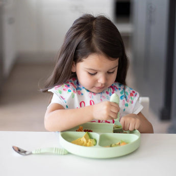A girl with long hair in a floral bib eats from a Bumkins Silicone Grip Dish: Sage with matching utensils at a white table.
