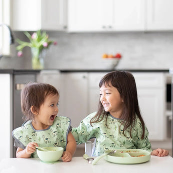 Two kids in matching bibs smile at each other while eating at a white table; one uses a Bumkins Silicone Grip Dish: Sage.