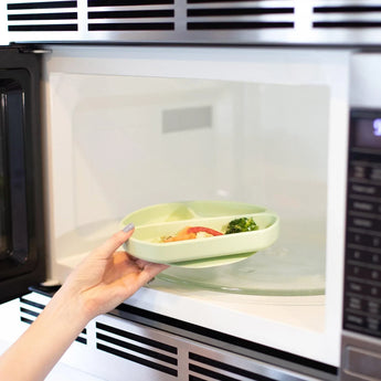 A person places a Bumkins Silicone Grip Dish: Sage with food into an open microwave.