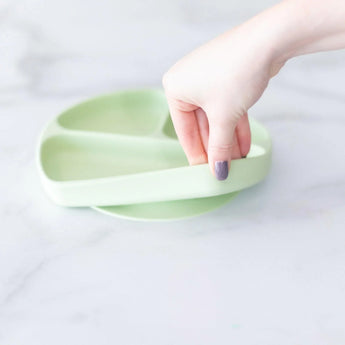A hand holds a Bumkins Silicone Grip Dish: Sage, demonstrating the suction cup bottom on a marble surface.