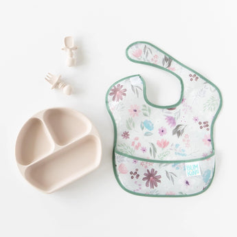 Bumkins Silicone Grip Dish: Sand sits next to matching toddler utensils and a floral bib on a white background.