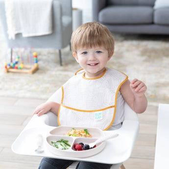 A toddler smiles in a high chair with food in Bumkins Silicone Grip Dish: Sand on the tray, next to matching utensils.