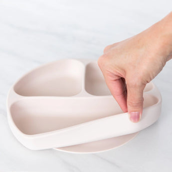 A hand holds a Bumkins Silicone Grip Dish: Sand, demonstrating the suction cup bottom on a marble surface.