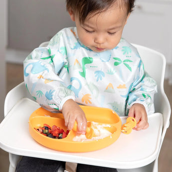 A toddler in a high chair wearing a dinosaur bib enjoys yogurt and fruit from a Bumkins Silicone Grip Dish: Tangerine with orange utensils.