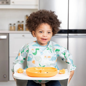 A curly-haired child in a dinosaur bib smiles in a high chair with a Bumkins Silicone Grip Dish: Tangerine and orange utensils on the tray.