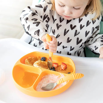 A toddler in a high chair wearing a black and white bib enjoys breakfast from a Bumkins Silicone Grip Dish: Tangerine with a fork.