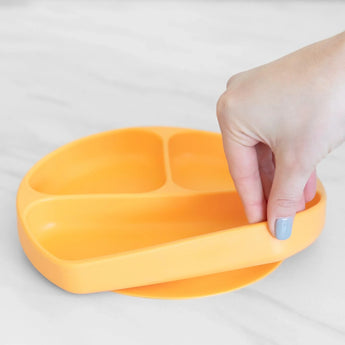 A hand holds a Bumkins Silicone Grip Dish: Tangerine, demonstrating the suction cup bottom on a marble surface.