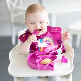 A baby in a high chair enjoys food from a Bumkins Silicone Grip Dish Special Edition: Unicorn with matching utensils, wearing a unicorn bib.
