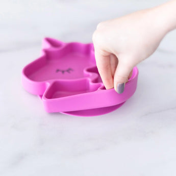 A hand holds a Bumkins Silicone Grip Dish Special Edition: Unicorn, demonstrating the suction cup bottom on a marble surface.