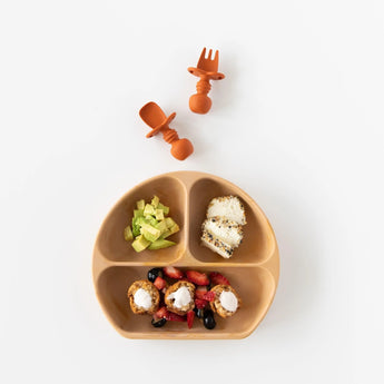 Bumkins Silicone Grip Dish: Wood Grain holds avocado, toast, and yogurt, with two orange toddler utensils above on a white background.