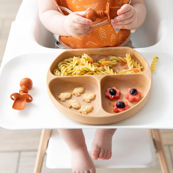 A baby in a highchair, wearing an orange bib, holds a spoon and enjoys pasta, crackers, and fruit from Bumkins Silicone Grip Dish: Wood Grain.