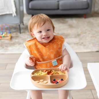 A toddler in a high chair wearing an orange bib enjoys food from Bumkins Silicone Grip Dish: Wood Grain.