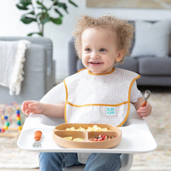 A smiling toddler in a high chair holds a spoon next to a Bumkins Silicone Grip Dish: Wood Grain filled with food.