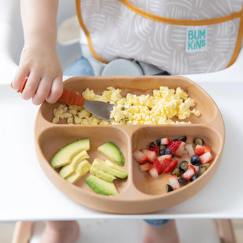 Close-up of a child in a high chair eating scrambled eggs, avocado, and fruit from Bumkins Silicone Grip Dish: Wood Grain with a fork.