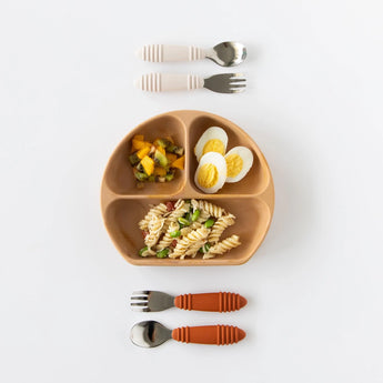 Bumkins Silicone Grip Dish: Wood Grain holds pasta, eggs, and fruit salad on a white background, with two sets of utensils above and below.