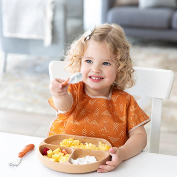 A child with curly hair holds a spoon over a divided plate on a white table, wearing an orange Bumkins Junior Bib.